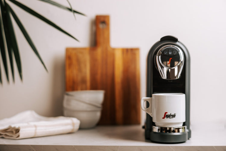 A coffee machine with a white cup is on a countertop, next to a cutting board, bowls, and a folded cloth.