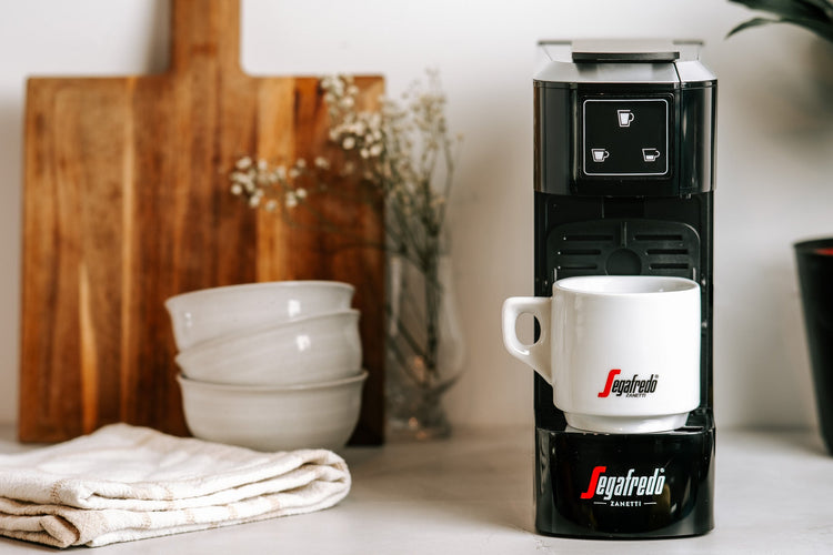 A coffee machine with a white cup is on a countertop, next to a cutting board, bowls, and a folded cloth.