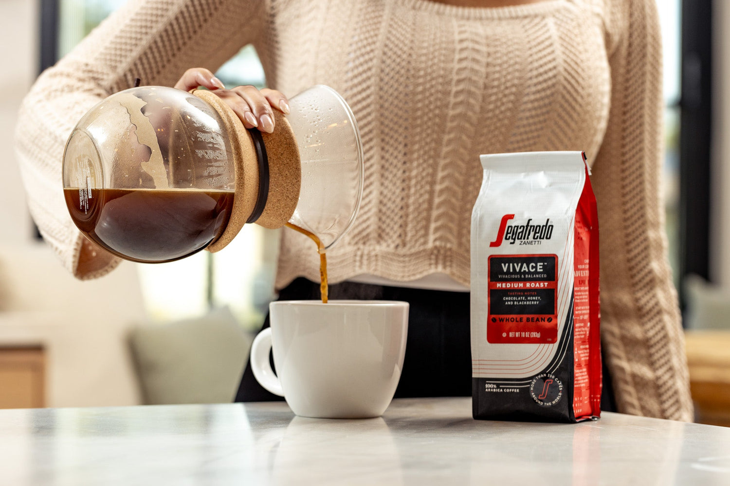 Person pours coffee from a Chemex into a mug, with a bag of Sagafredo Vivace coffee beans placed nearby on a countertop.