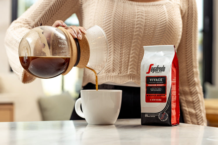 Person pours coffee from a Chemex into a mug, with a bag of Sagafredo Vivace coffee beans placed nearby on a countertop.