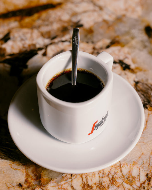 A Segafredo Zanetti Segafredo Cup & Saucer Set, filled with black coffee and a metal spoon inside, rests on a marbled surface.
