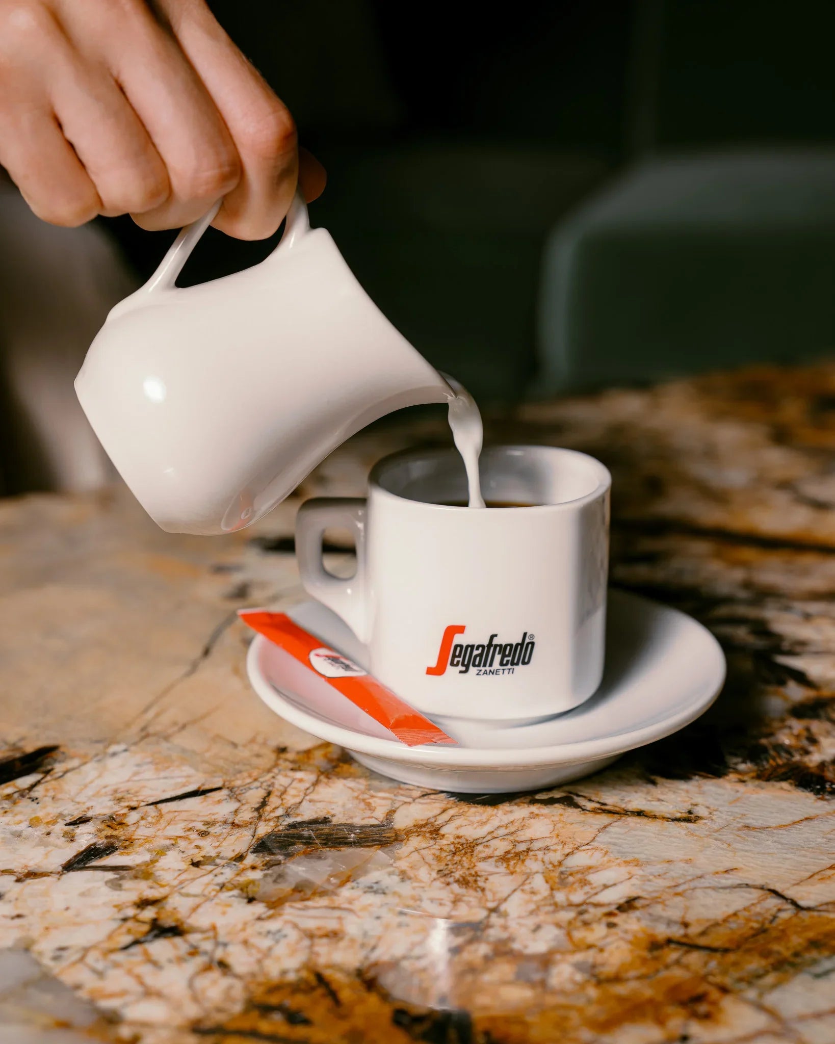 Milk is poured from a white creamer into a Segafredo Zanetti Cup & Saucer Set, featuring the brand’s ceramic espresso cup with saucer and red packet, all arranged on a marble table.