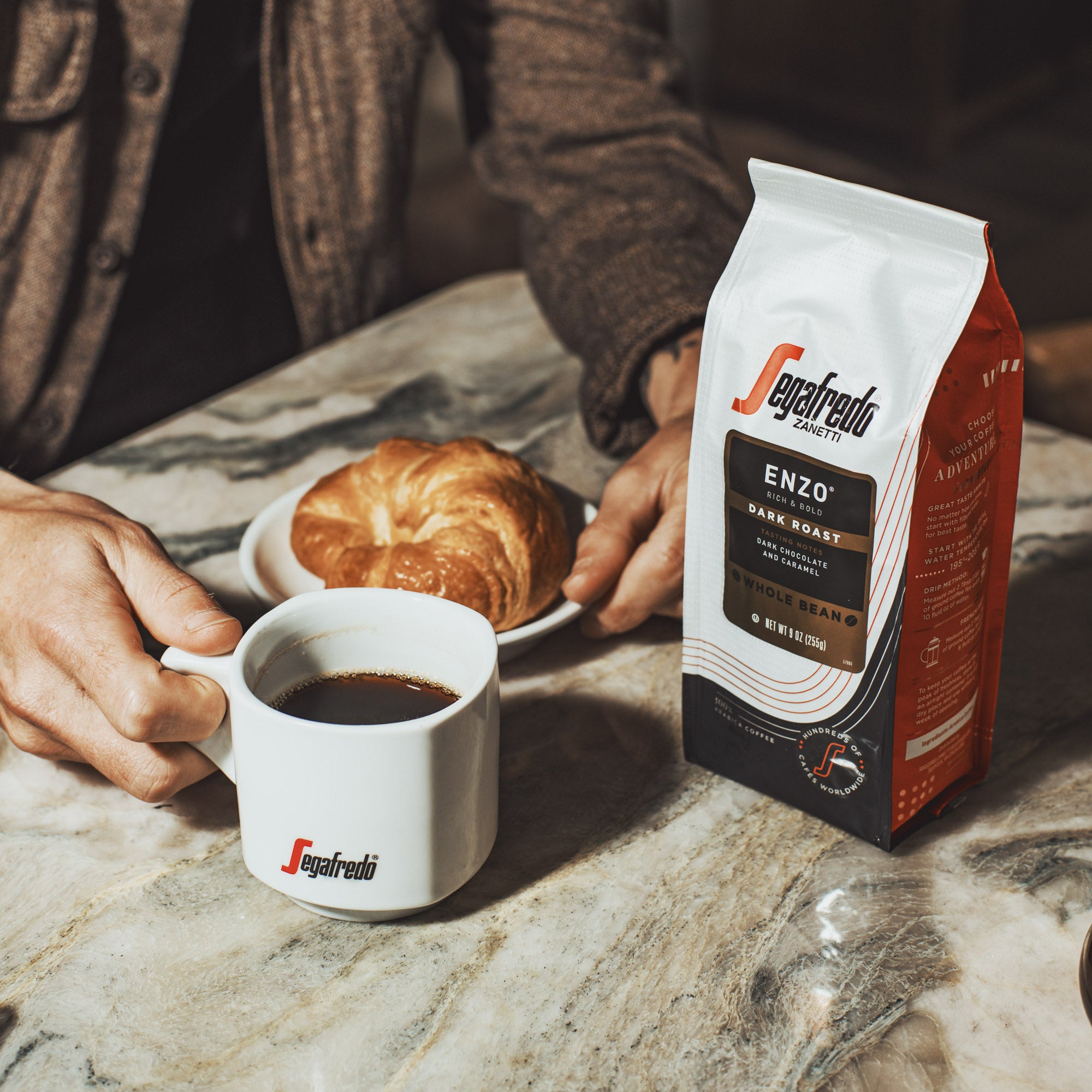 A person holds a mug of coffee beside a croissant and a bag of Segafredo Zanetti® Coffee Enzo Dark Roast whole bean coffee on a marble table.
