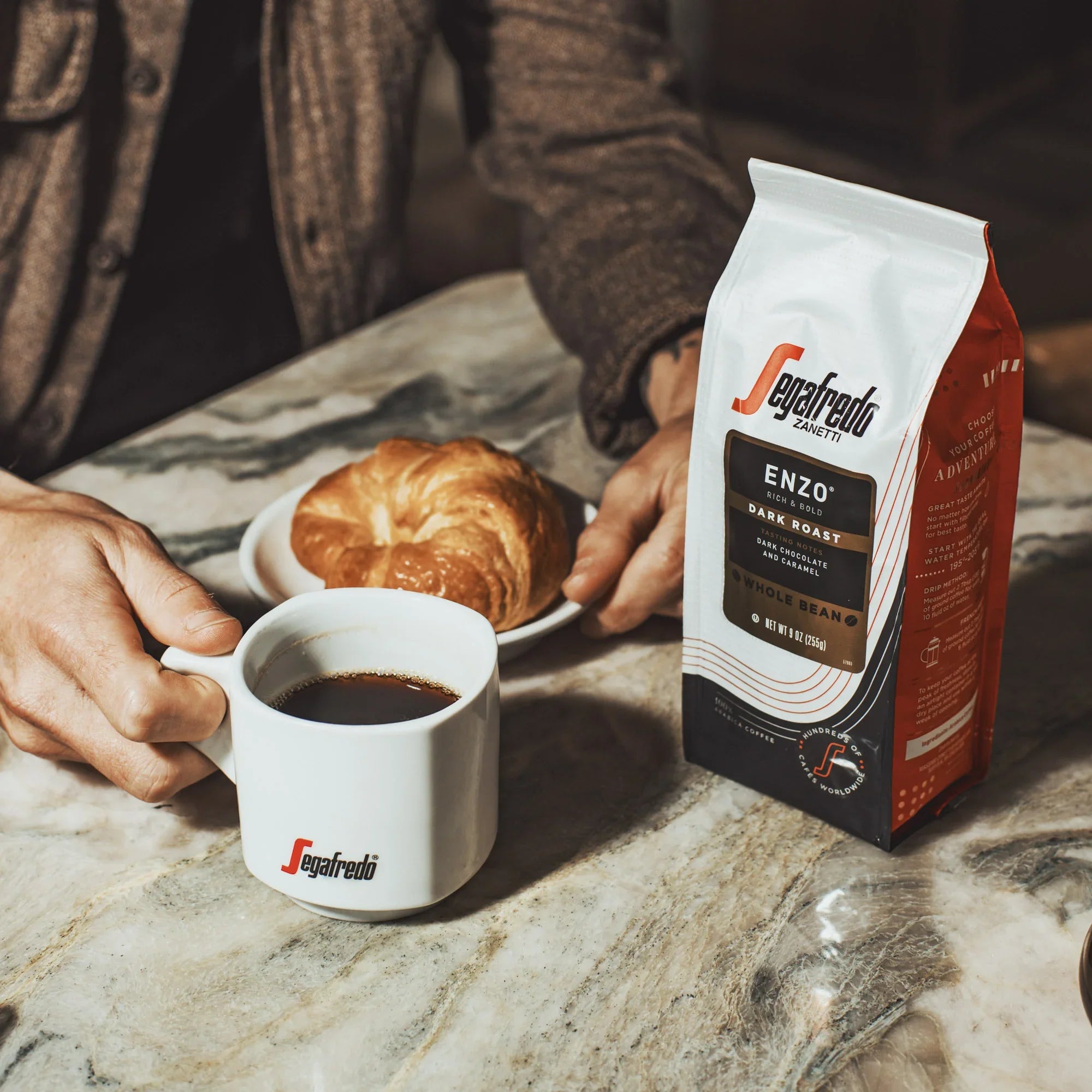 A person holds a mug of coffee beside a croissant and a bag of Segafredo Zanetti® Coffee Enzo Dark Roast whole bean coffee on a marble table.