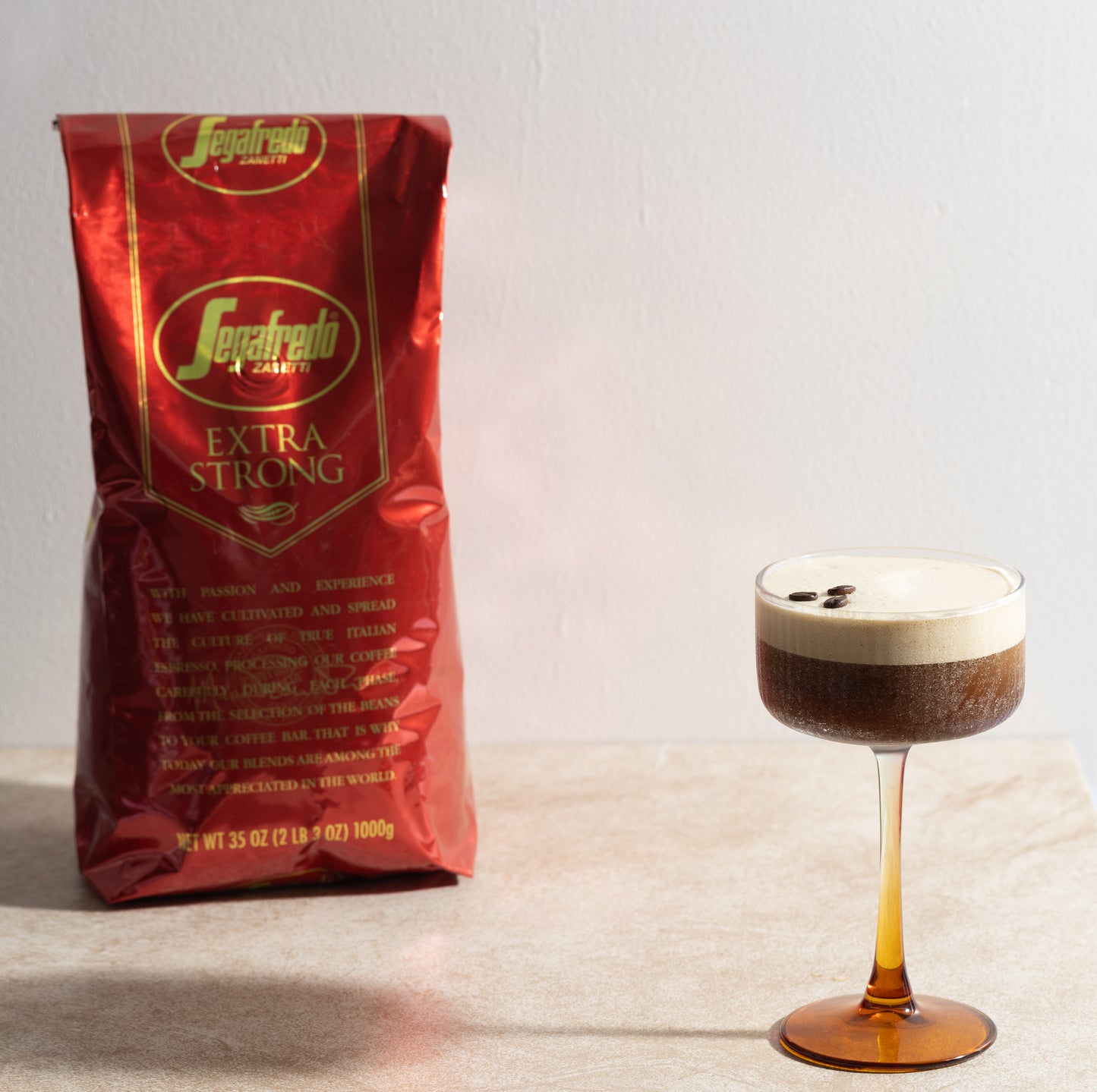 A red bag of Segafredo Zanetti® Coffee Extra Strong Whole Bean Espresso stands behind a cocktail glass filled with a frothy espresso topped with three coffee beans and rich crema, highlighting this dark roast.