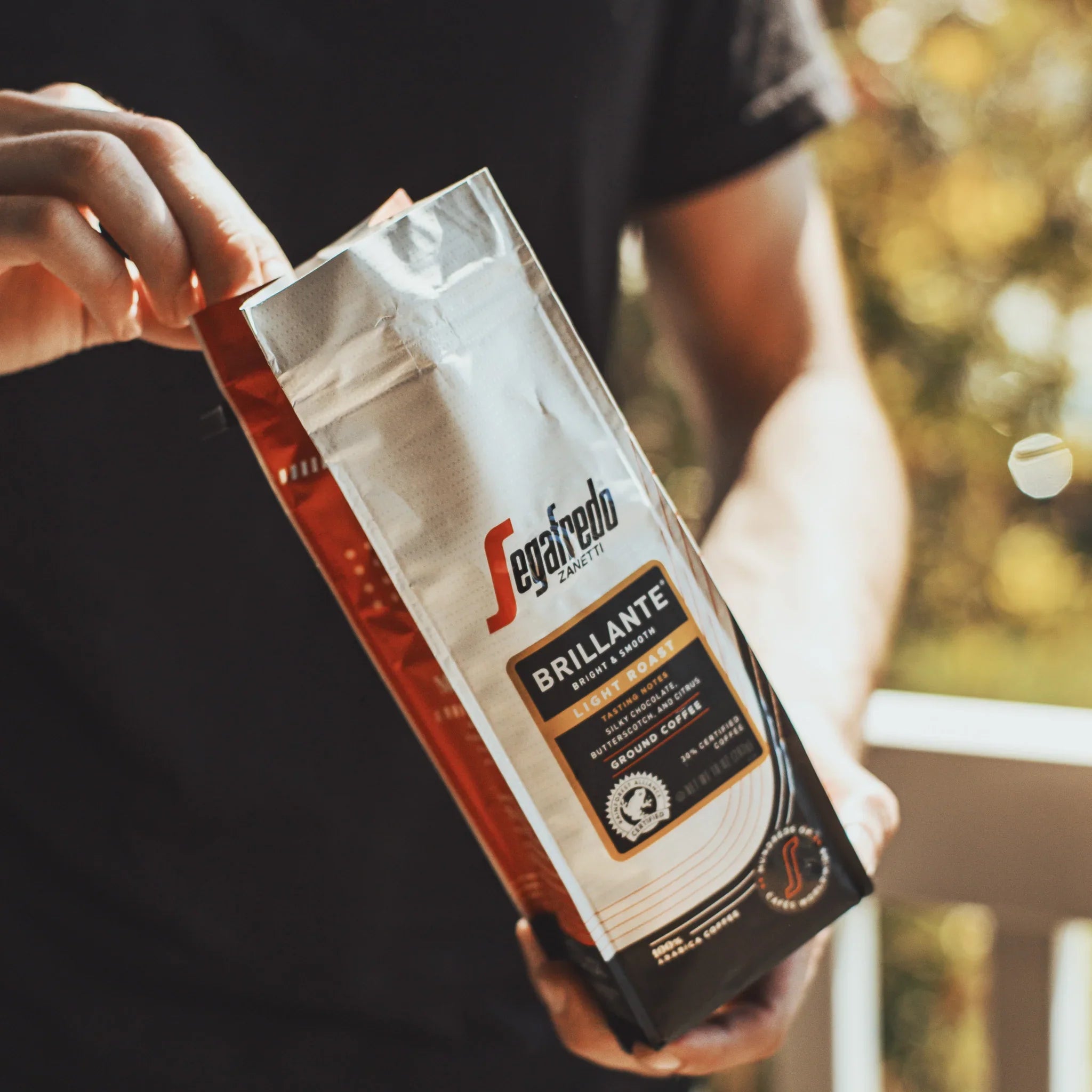 Standing outdoors, someone opens a bag of Brillante Light Roast by Segafredo Zanetti® Coffee, made from Colombian Arabica beans.