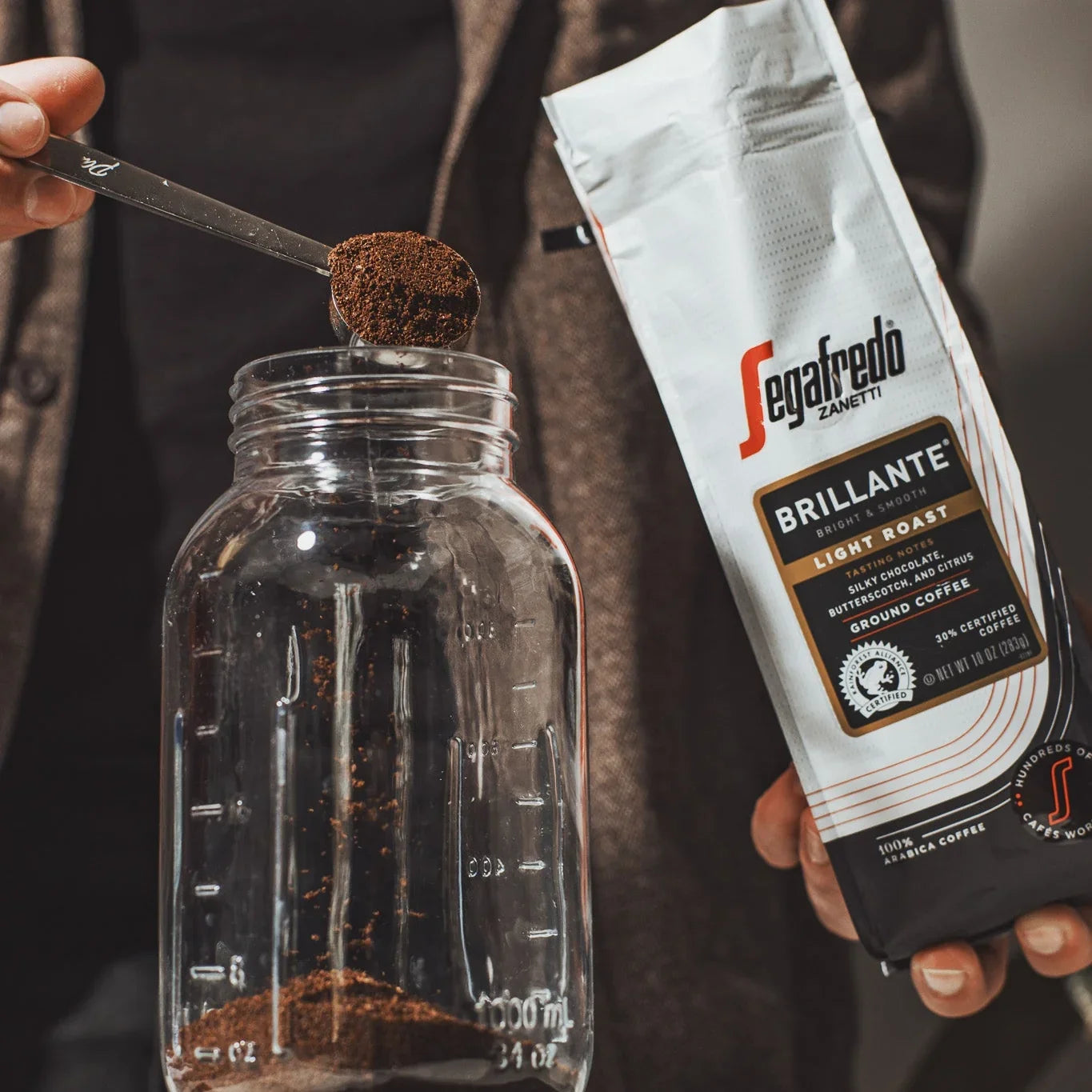 Scooping Brillante Light Roast by Segafredo Zanetti® Coffee from its bag into a glass jar—ideal for fans of lightly roasted coffee or a smooth cold brew.