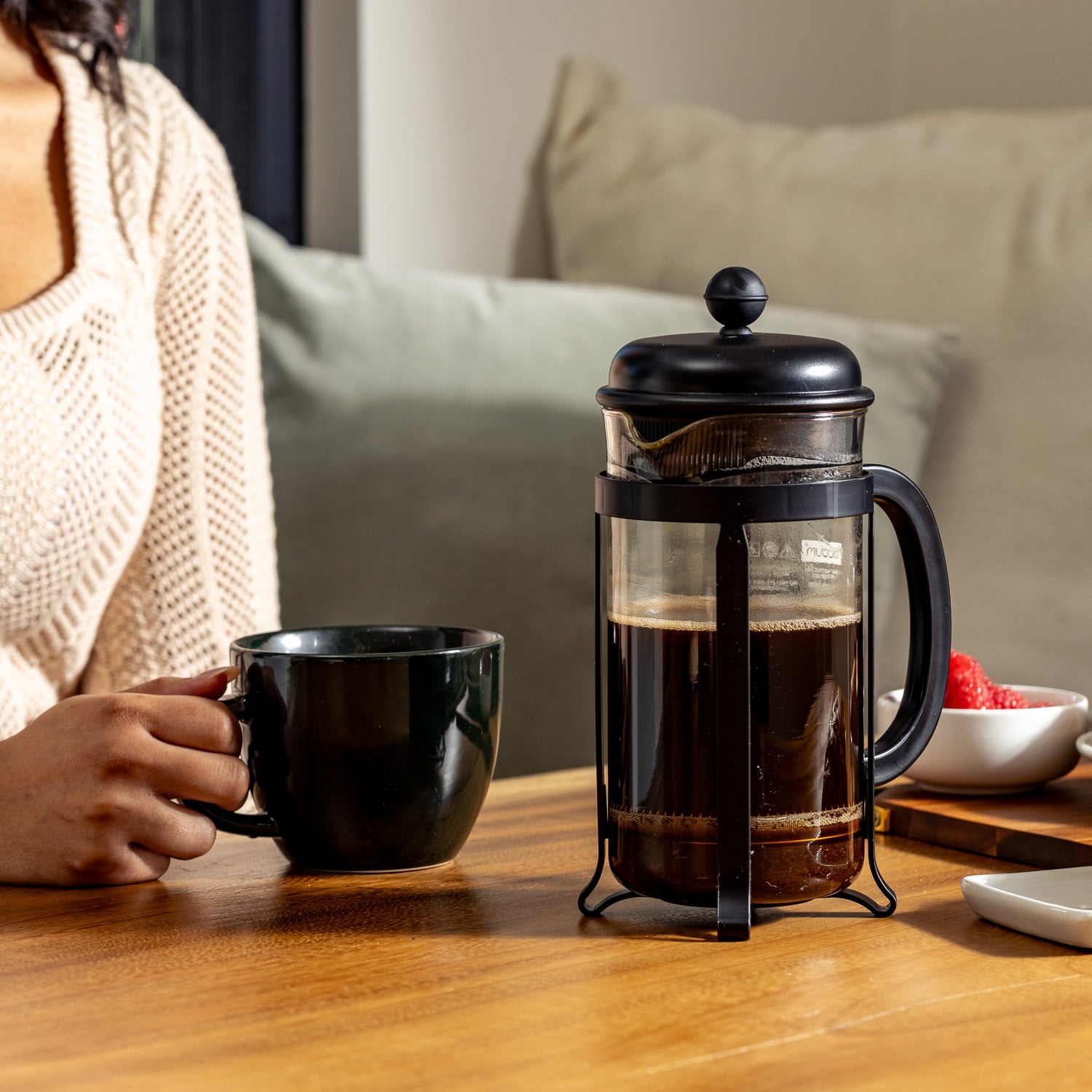 A person holds a black mug next to a French press with Segafredo Zanetti® Coffee's Vivace Medium Roast, featuring hints of chocolate, on a wooden table, alongside a bowl of strawberries.