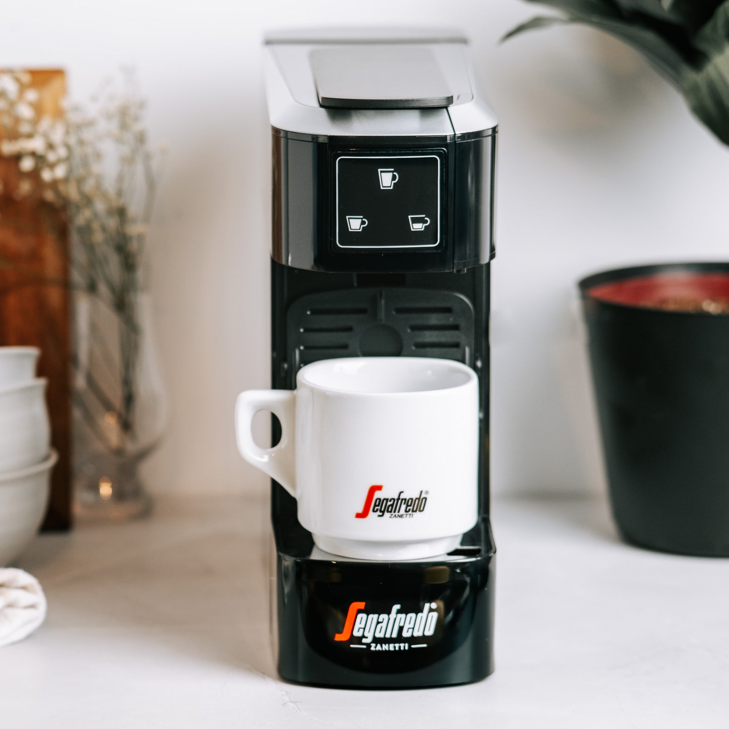 A Sky Extra Espresso Capsule Machine by Segafredo Zanetti® Coffee sits on a white countertop with a matching Segafredo mug, near a plant and some cups.
