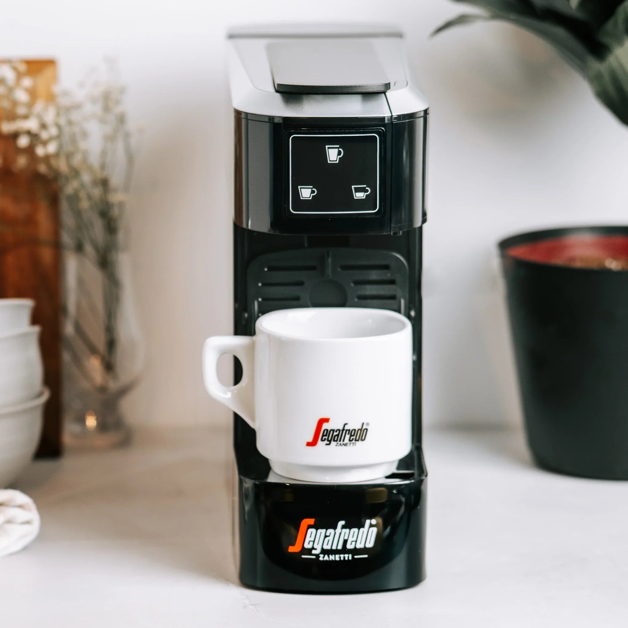 A Sky Extra Espresso Capsule Machine by Segafredo Zanetti® Coffee sits on a white countertop with a matching Segafredo mug, near a plant and some cups.