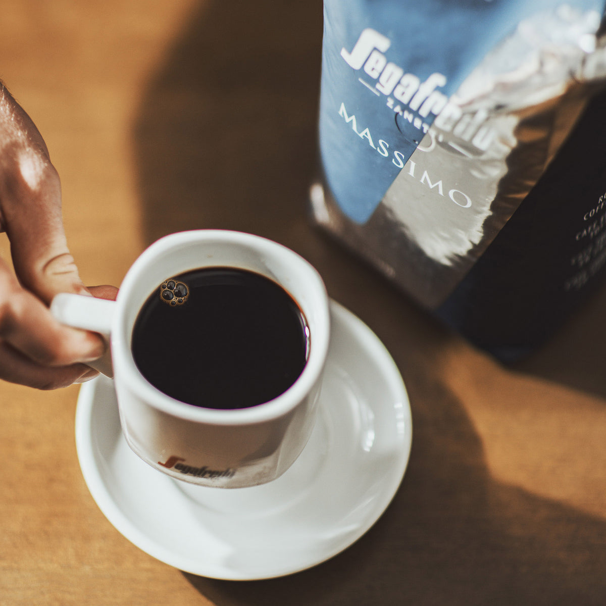 A hand holding a white cup of black espresso next to a bag of Segafredo Massimo Whole Bean Espresso.