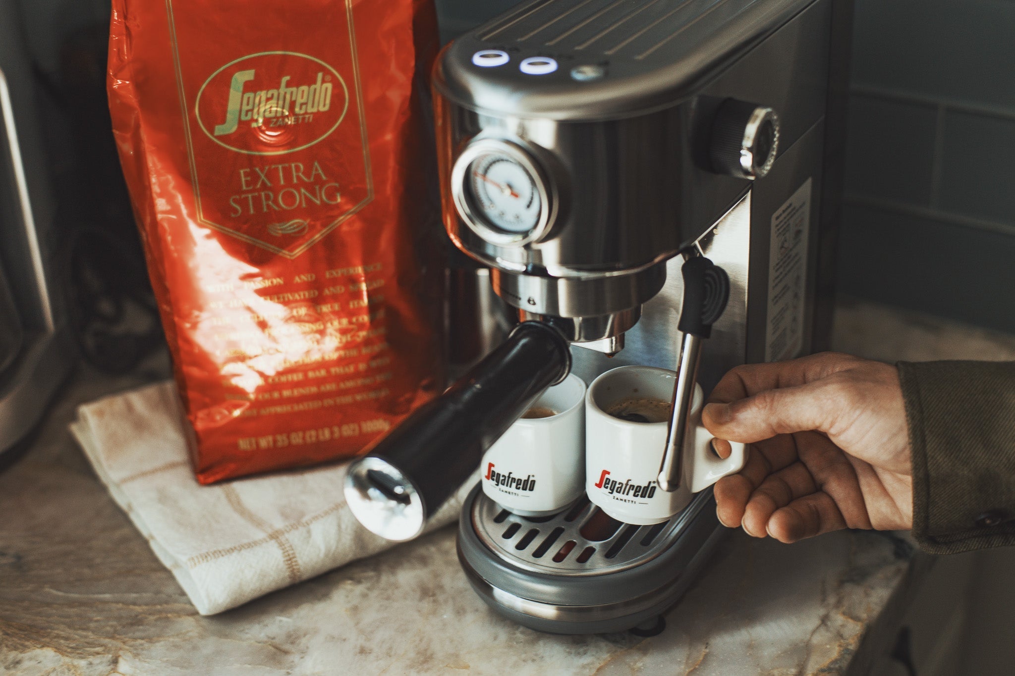 A person uses a stainless steel espresso machine to brew coffee into two small Segafredo cups, with a red bag of Segafredo Extra Strong coffee beside the machine.