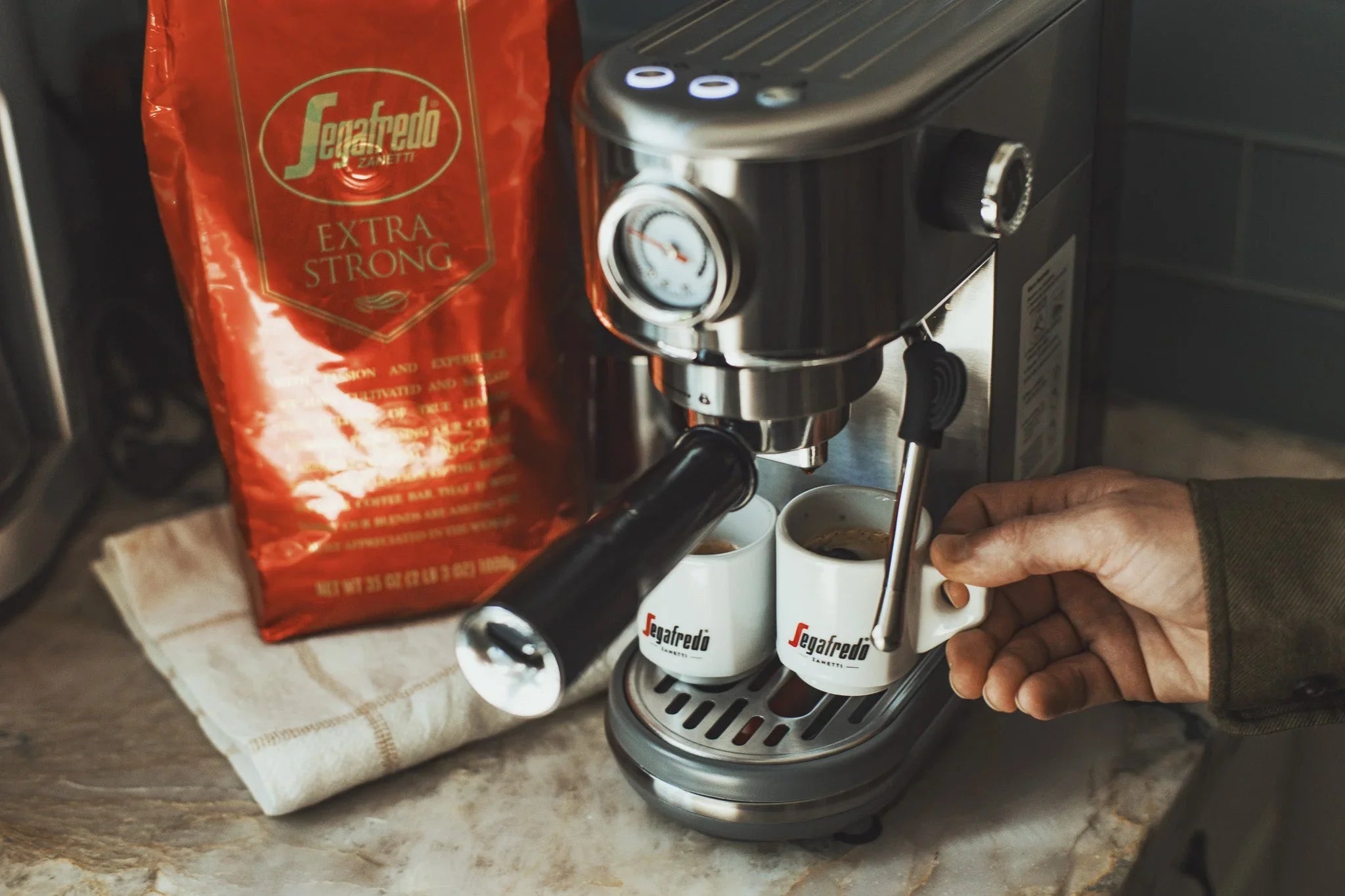 A person uses a stainless steel espresso machine to brew coffee into two small Segafredo cups, with a red bag of Segafredo Extra Strong coffee beside the machine.