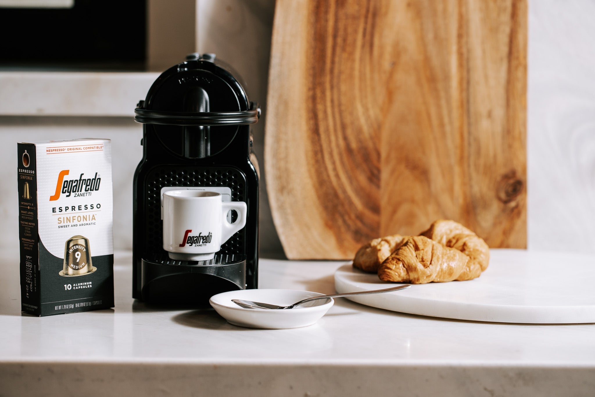 A Segafredo espresso machine with a cup, a box of Segafredo Sinfonia espresso pods, a spoon, and two croissants on a kitchen counter.