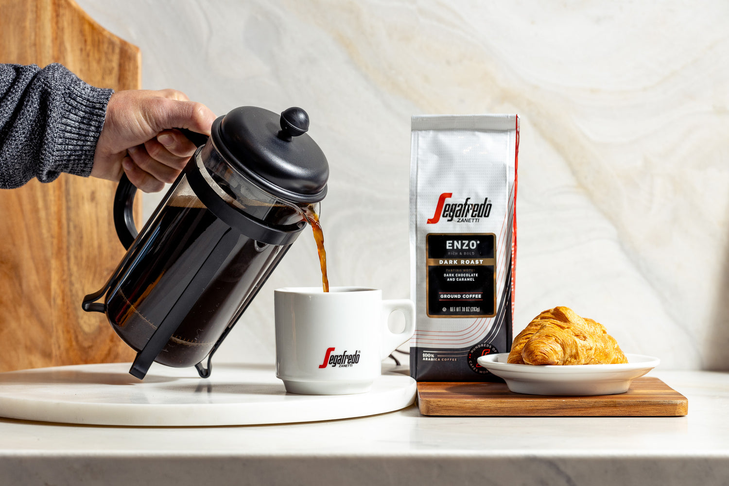 A person pours coffee from a French press into a white mug. A bag of Segafredo Enzo Dark Roast coffee beans and a croissant on a plate are set beside it.