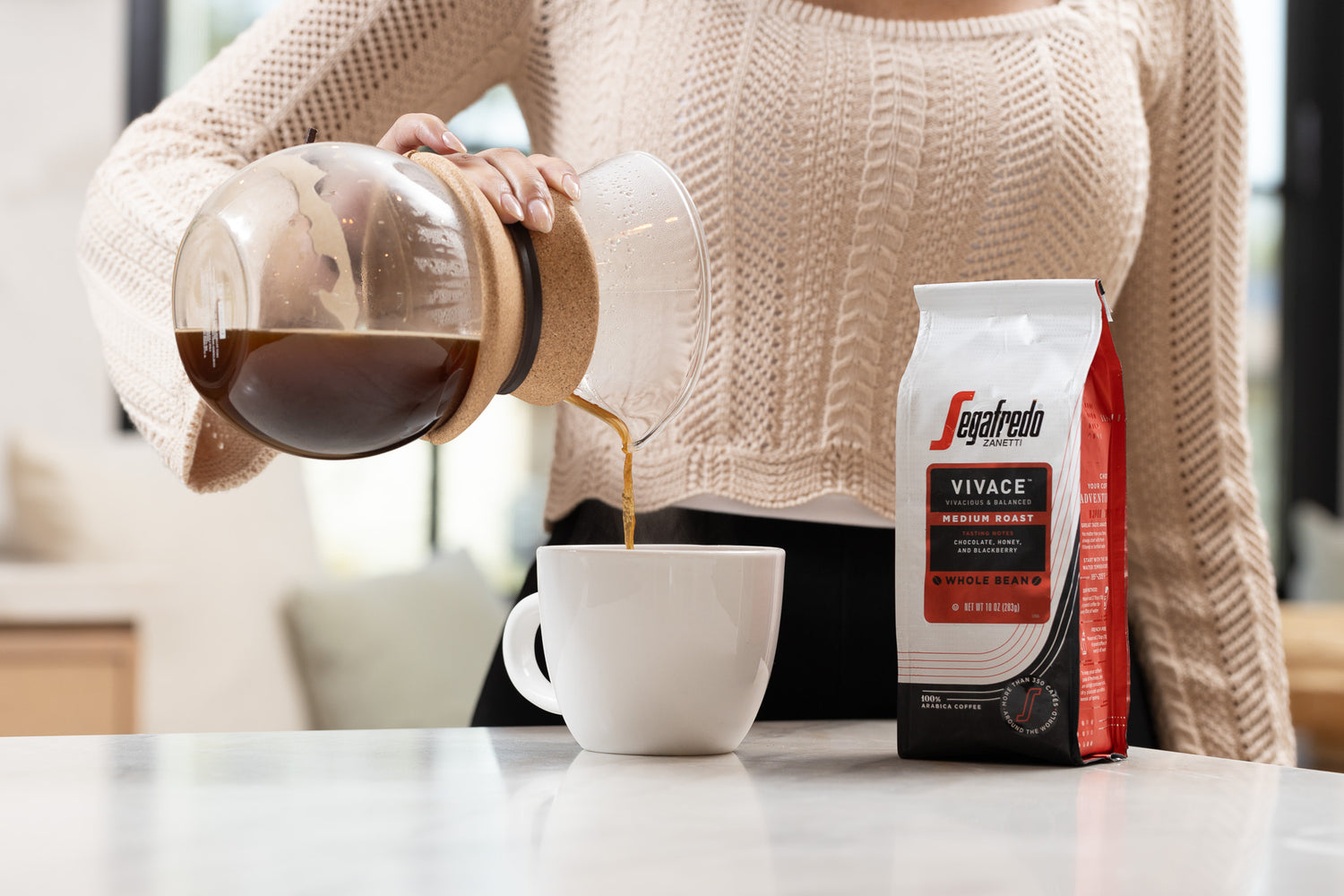 Person pouring coffee from a carafe into a white mug next to a bag of Segafredo Vivace Medium Roast whole bean coffee on a countertop.