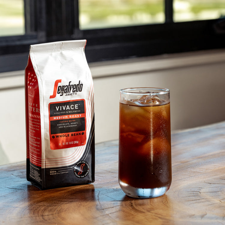 A bag of Segafredo Zanetti Vivace medium roast whole bean coffee is next to a glass of iced coffee on a wooden surface.