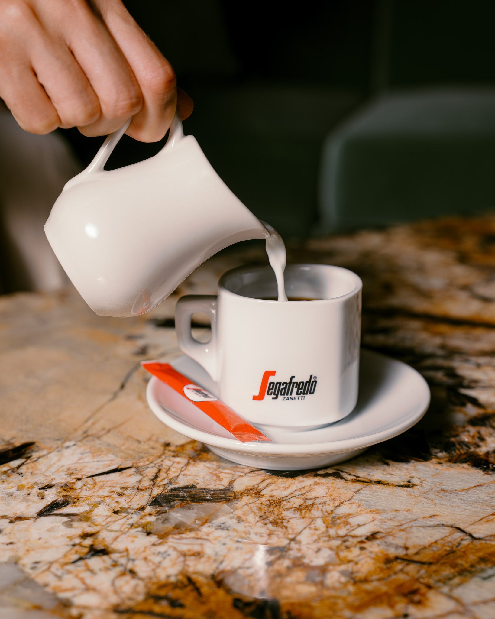 Milk is poured from a white creamer into a Segafredo Zanetti Cup & Saucer Set, featuring the brand’s ceramic espresso cup with saucer and red packet, all arranged on a marble table.