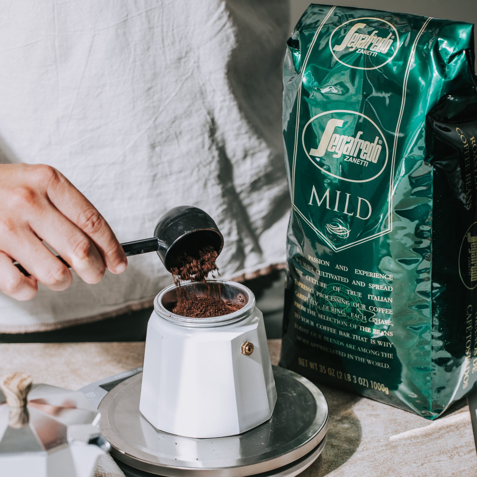 A person scoops Segafredo Zanetti® Coffee Mild Whole Bean Espresso into a moka pot on a scale.