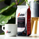 Load image into Gallery viewer, A bag of Segafredo Zanetti® Coffee Crescendo Medium Roast Ground Coffee is placed next to a white coffee mug with the Segafredo logo. Green foliage is visible in the background, hinting at the earthy undertones of this Italian coffee style.

