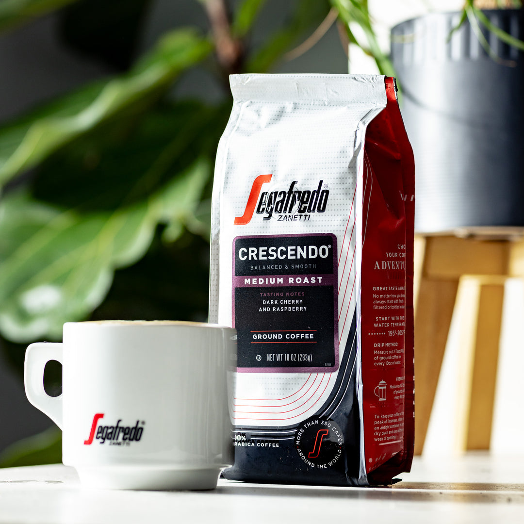 A bag of Segafredo Zanetti® Coffee Crescendo Medium Roast Ground Coffee is placed next to a white coffee mug with the Segafredo logo. Green foliage is visible in the background, hinting at the earthy undertones of this Italian coffee style.