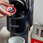 Load image into Gallery viewer, A hand places a single-serve coffee pod labeled &quot;Vivace Medium Roast Keurig K-Cup® Pods&quot; from Segafredo Zanetti® Coffee, a rich Latin American blend, into a Keurig coffee machine, with a box and white mug nearby, emphasizing its KEURIG 2.0 COMPATIBLE nature.
