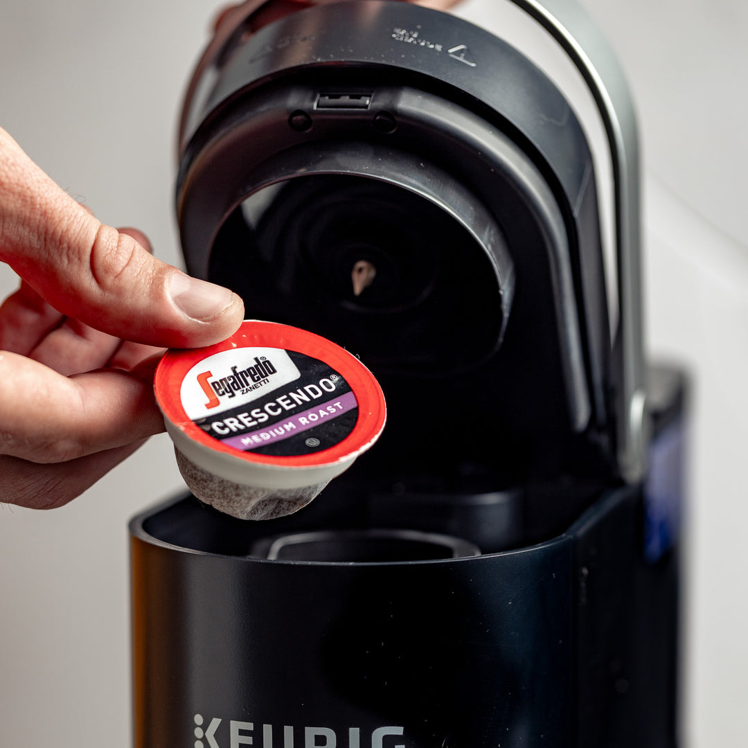 A hand placing a Segafredo Zanetti® Coffee Crescendo Medium Roast Keurig K-Cup® Pod in a Keurig coffee machine, capturing the essence of Italian coffee style with its low fruit tones.