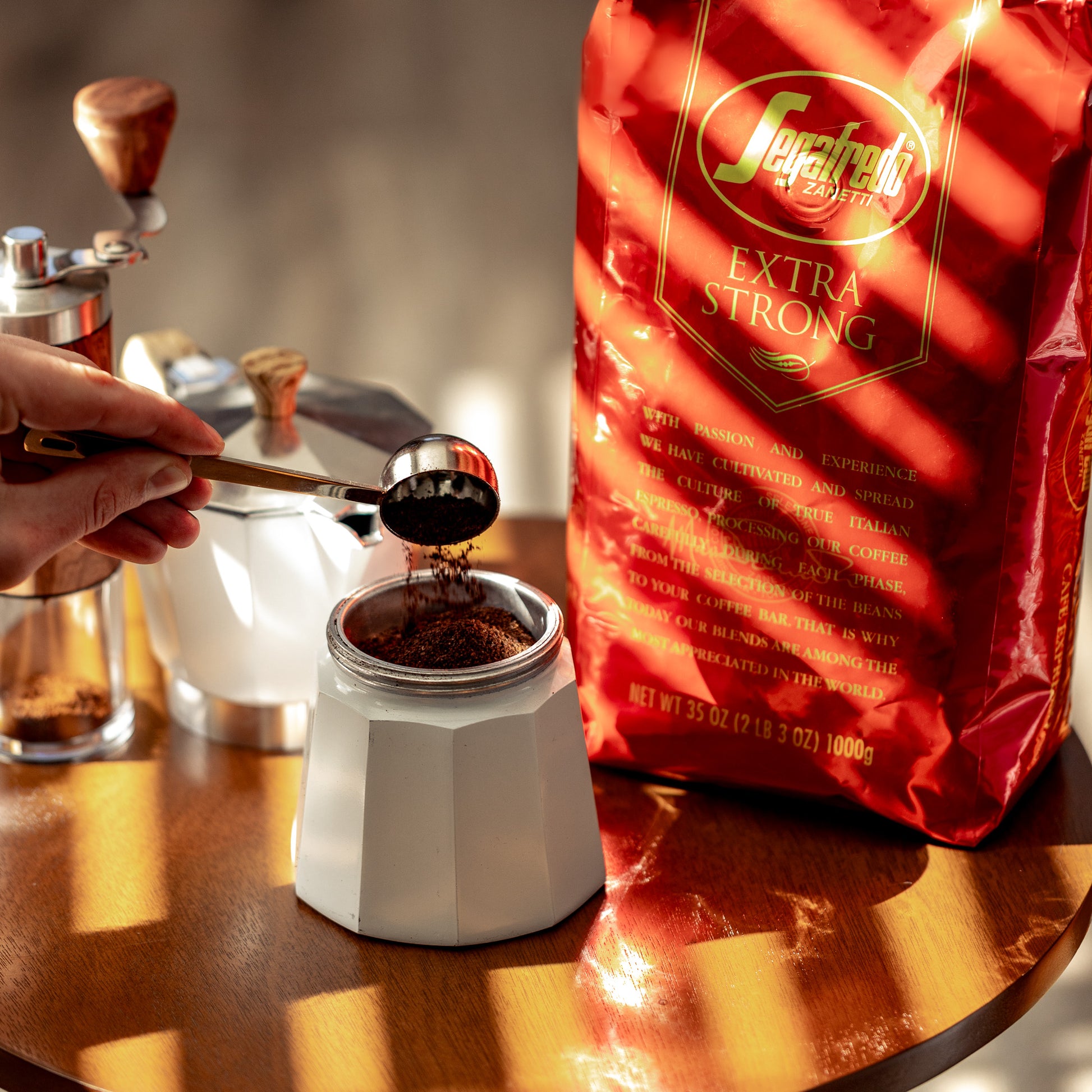 A person fills a moka pot with ground coffee using a metal scoop. On the table is a red bag of Segafredo Zanetti® Coffee Extra Strong Whole Bean Espresso.