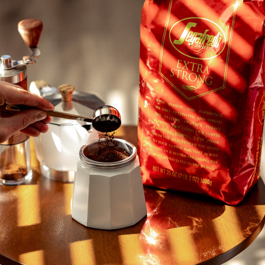 A person fills a moka pot with ground coffee using a metal scoop. On the table is a red bag of Segafredo Zanetti® Coffee Extra Strong Whole Bean Espresso.