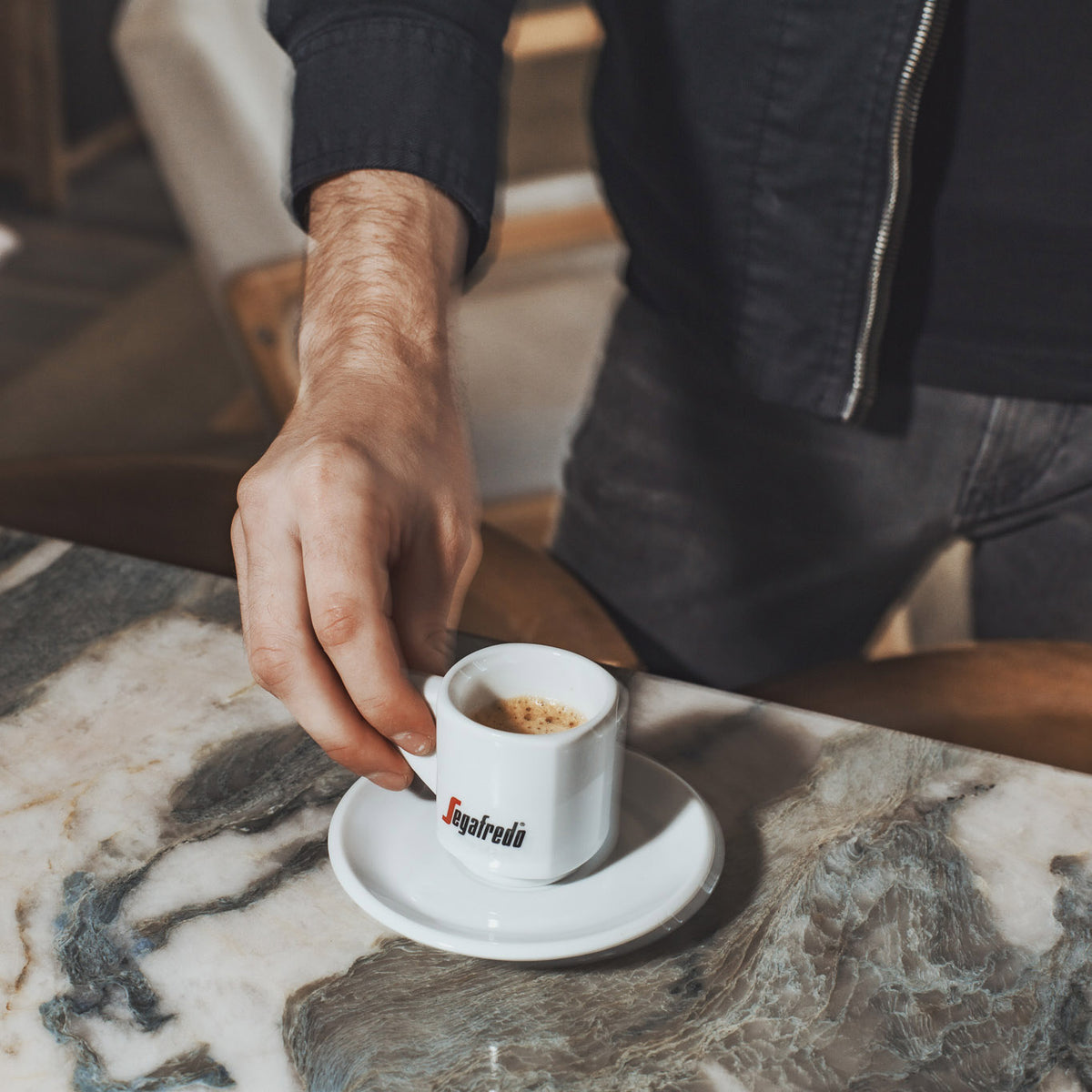A person holding a small white Segafredo espresso cup on a saucer, placed on a marble table, enjoying the rich aroma of Italian espresso brewed from Segafredo Massimo Whole Bean Espresso.