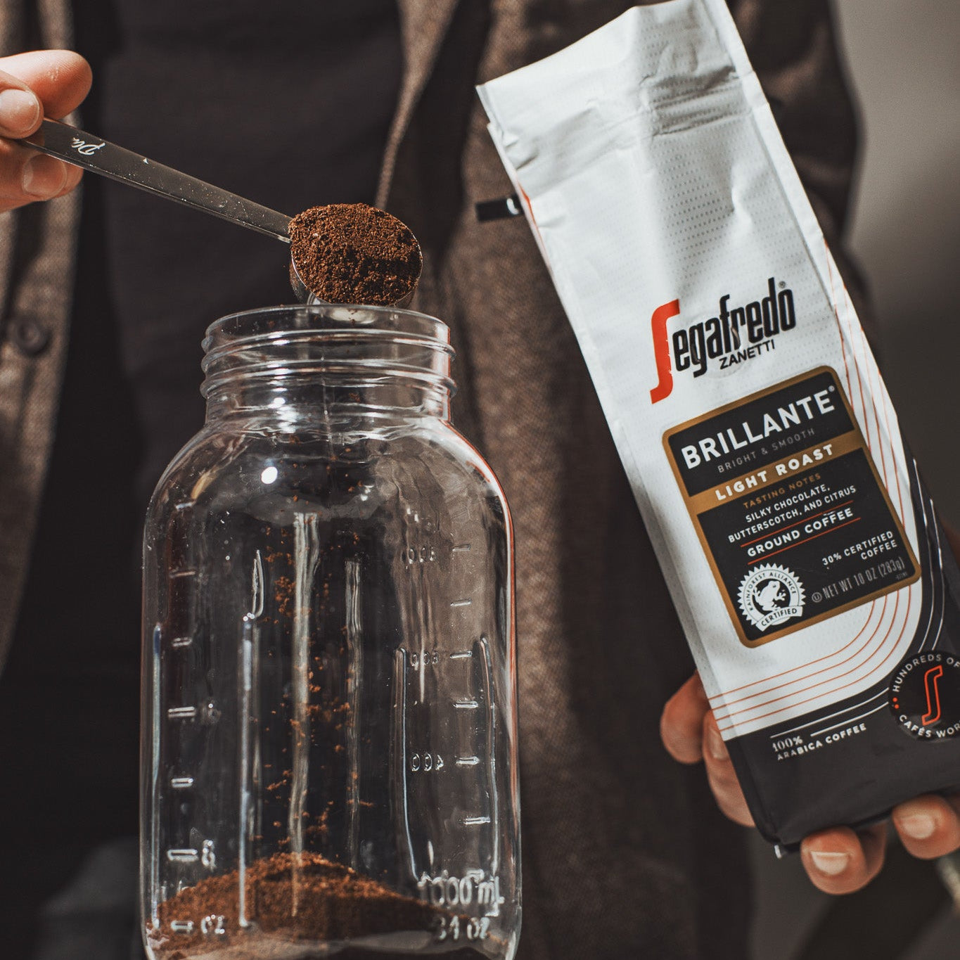 Scooping Brillante Light Roast by Segafredo Zanetti® Coffee from its bag into a glass jar—ideal for fans of lightly roasted coffee or a smooth cold brew.
