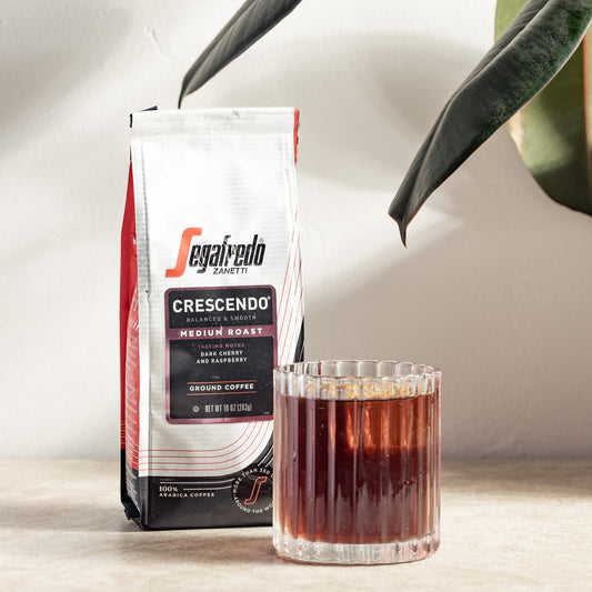 A bag of Segafredo Zanetti® Coffee Crescendo Medium Roast sits beside a glass of brewed coffee on a light surface, with green plant leaves partially visible above.