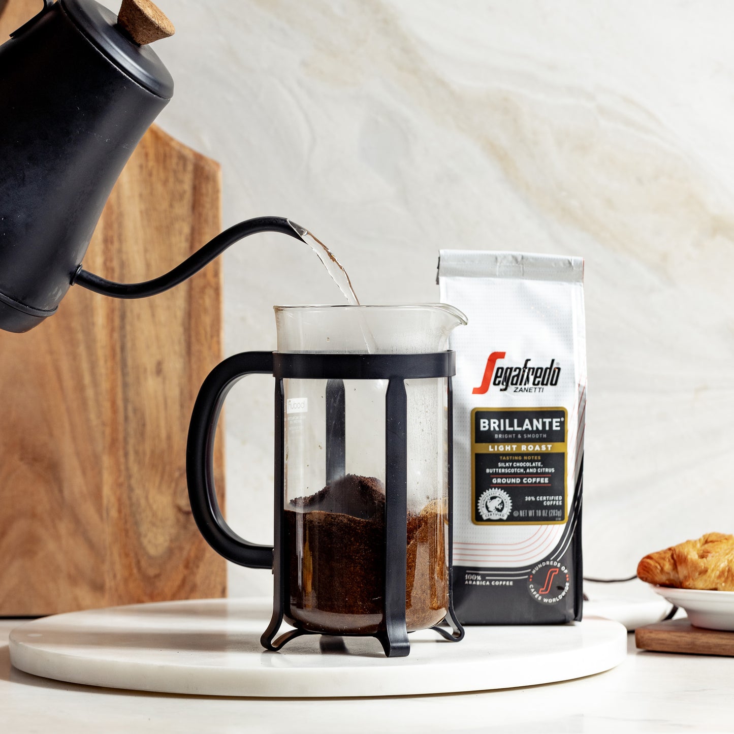 A black kettle pouring hot water into a French press with coffee grounds beside a package of Segafredo Zanetti® Coffee Brillante Light Roast Ground Coffee and a croissant on a plate.