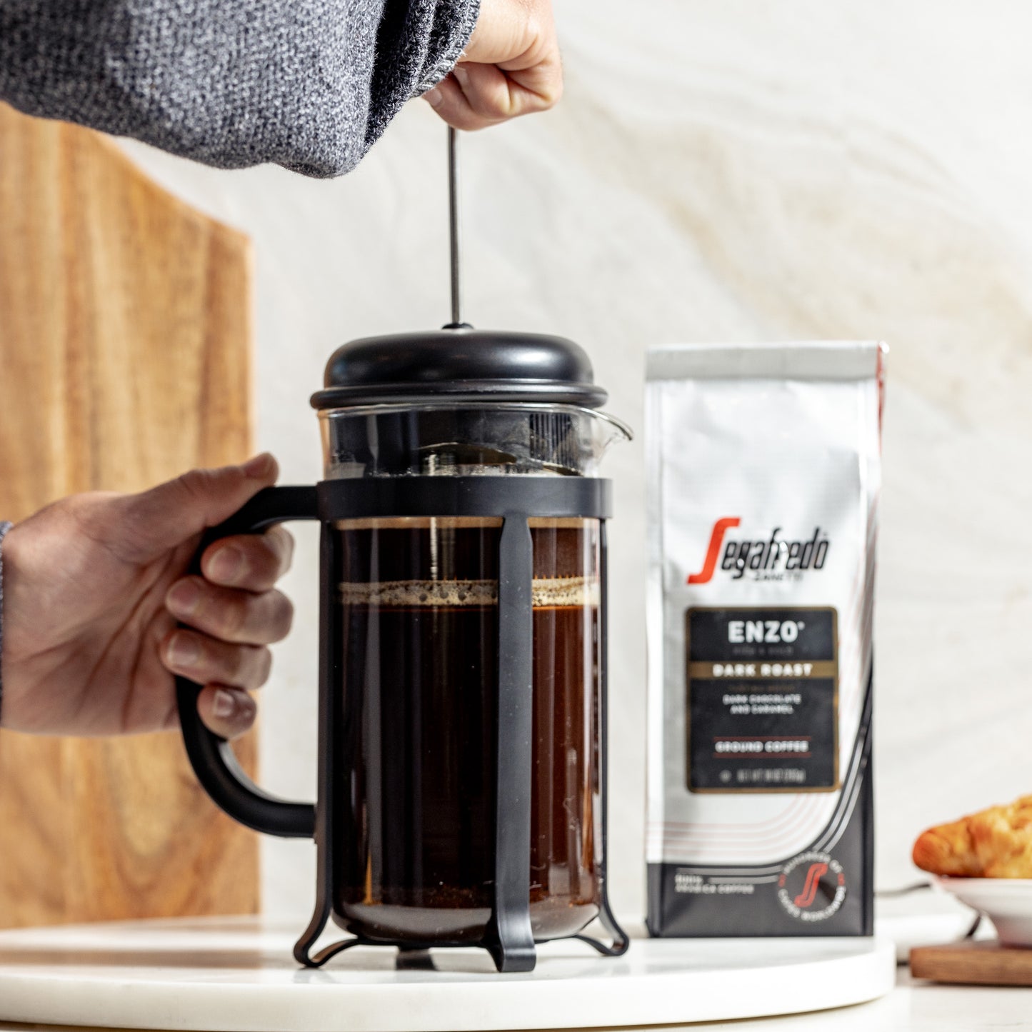 A close-up of a person's hand pressing down the plunger of a French press. In the background, there is a bag of Segafredo Zanetti® Coffee Enzo Dark Roast Ground Coffee, a rich craft blend known for its dark roasted intensity.