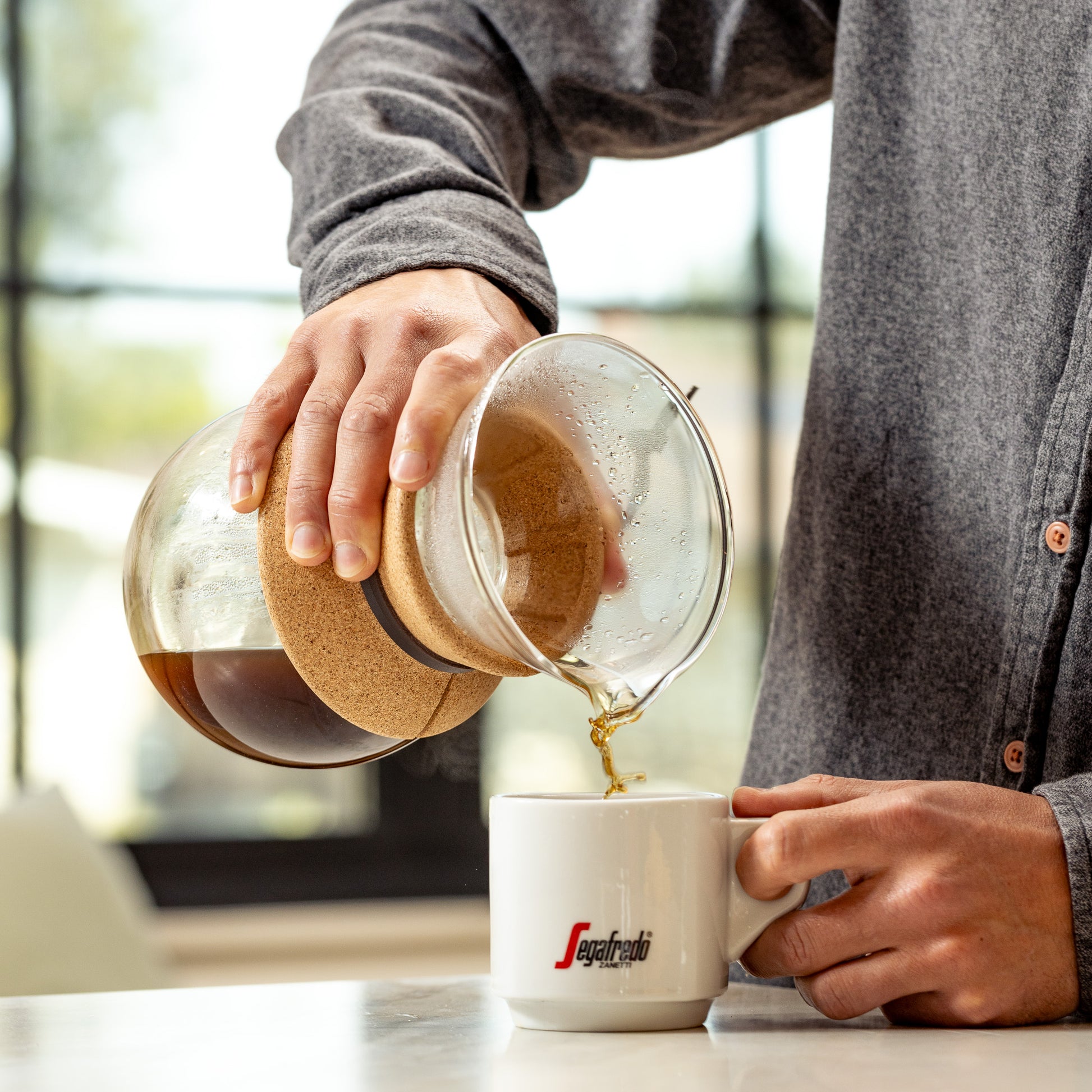 A person in a grey shirt pours Enzo Dark Roast Whole Bean Coffee from a glass carafe into a white mug with a red "Segafredo" logo in a bright room.