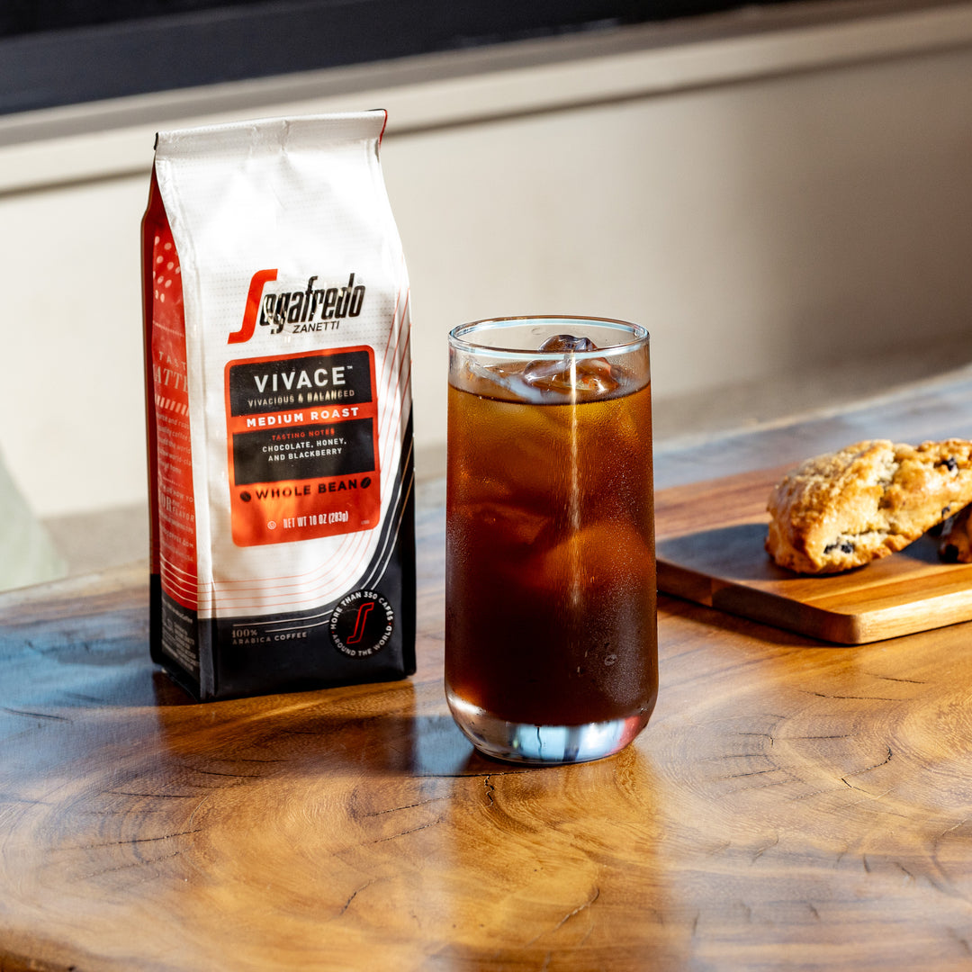 A glass of iced coffee sits on a wooden table beside a package of Vivace Medium Roast Whole Bean Coffee from Segafredo Zanetti® Coffee, promising a full-bodied cup with rich chocolate and caramel notes. A pastry rests on a wooden board nearby.