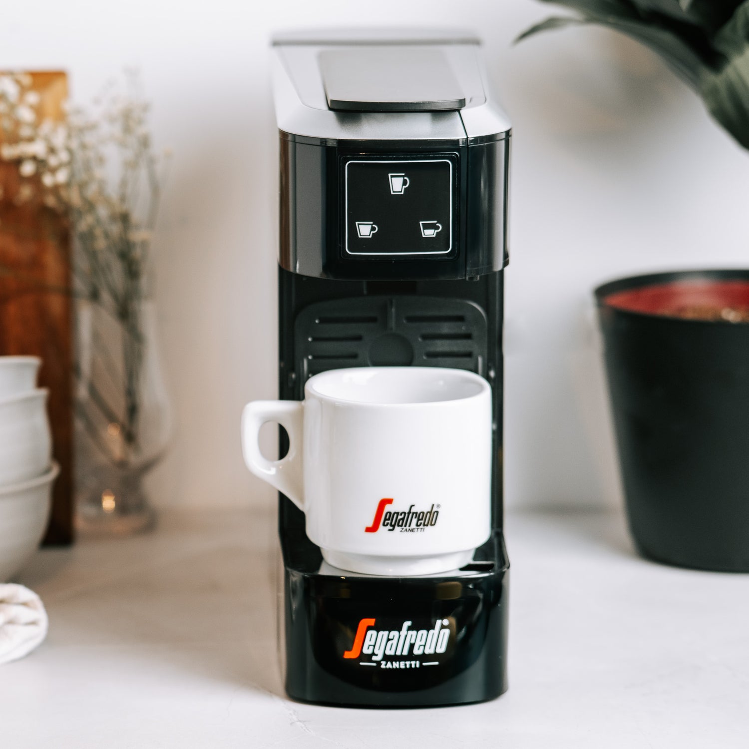 A Sky Extra Espresso Capsule Machine by Segafredo Zanetti® Coffee sits on a white countertop with a matching Segafredo mug, near a plant and some cups.